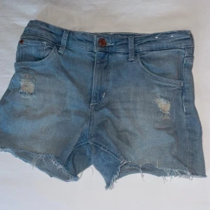 Ljusblå jeansshorts med fransar - Säljer ett par ljusblå jeansshorts från Bm med slitna detaljer och fransig nederkant. Klassisk femficksmodell med knapp och dragkedja framtill. Perfekta för varma dagar. Mid waist