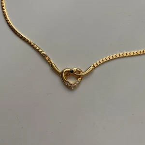 Guldfärgat halsband med hjärthänge - Snyggt guldfärgat halsband med ett hjärta i mitten, dekorerat med små glittrande stenar och en mörk sten i centrum. Kedjan är platt och glänsande, perfekt för att ge en elegant touch till din outfit.