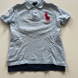  pikétröja från Polo Ralph Lauren - Säljer en ljusblå pikétröja från Polo Ralph Lauren med stor röd logga på bröstet och vit broderad siffra på ärmen. Baksidan är mörkblå med en stor vit siffra. Klassisk krage och knappar framtill. För barn