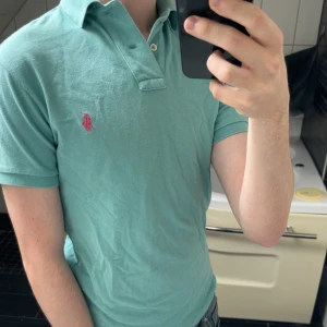 Mintgrön pikétröja från Ralph Lauren - Snygg mintgrön pikétröja från Ralph Lauren med klassisk krage och knappar. Tröjan har korta ärmar och den ikoniska rosa loggan broderad på bröstet. Perfekt för en avslappnad och stilren look.