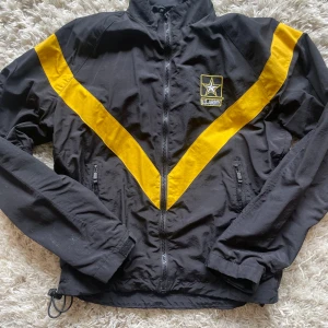 Svart och gul vindjacka från US Army - Svart vindjacka med gula V-formade detaljer och broderad US Army-logga på bröstet. Jackan har dragkedja framtill och två fickor med dragkedja. 