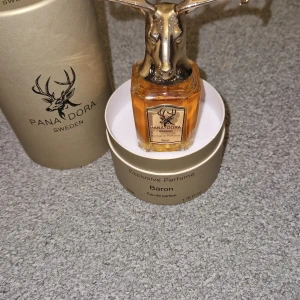 Pana Dora Baron Eau de Parfum 50ml - Exklusiv parfym från Pana Dora, Baron, 50ml. Flaskan är genomskinlig med guldiga detaljer och har ett unikt lock i form av ett hjorthuvud i metall. Parfymen kommer i en lyxig guldfärgad förpackning. Doften är Eau de Parfum och tillverkad i Sverige.
