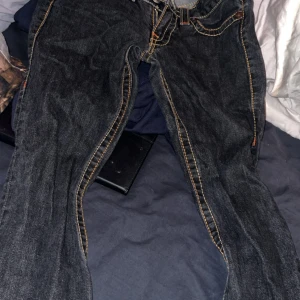 True Religion jeans med färgglada sömmar - Säljer ett par mörkblå True Religion jeans med ikoniska färgglada kontrastsömmar och snygga fickdetaljer bak. Klassisk femficksmodell med dragkedja och knappar framtill. Jeansen är i denim och har en unik look med broderade fickor.