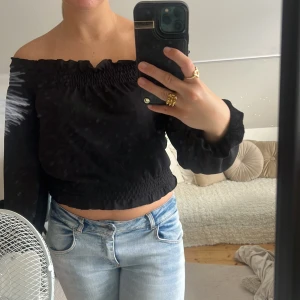 Svart offshoulder topp med smock - Trendig svart offshoulder topp med smockdetaljer upptill och nedtill. Långärmad och croppad modell som sitter snyggt över axlarna. Perfekt att styla med jeans eller kjol för en cool look.