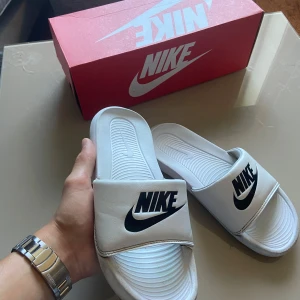 Vita Nike tofflor med svart logo - Snygga vita Nike sandaler med bred rem och stor svart Nike-logga framtill. Skorna har öppen tå och platt sula, perfekt för chill dagar. Tillverkade i syntetmaterial med bekväm innersula och klassisk design.