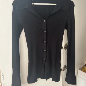 Svart ribbad kofta med krage - Säljer en svart ribbad kofta med lång ärm och snygg krage. Koftan har knappar hela vägen framtill och är figurnära i modellen. Perfekt att styla med jeans eller kjol för en clean look.