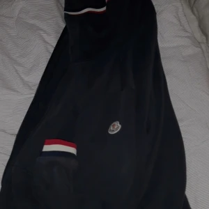 Svart Moncler hoodie - Svart hoodie från Moncler i storlek L. Passar M. Jackan har Moncler-logga på bröstet och coola ribbade muddar med röd, vit och marinblå rand. Perfekt för dig som vill ha en stilren och sportig look. Köp via plick! Nfc scan!