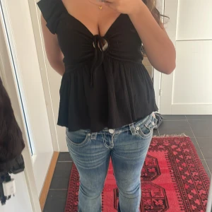 Svart blus med knyt och v-ringning - Supersöt svart blus med djup v-ringning och knyt framtill genom en rund ringdetalj. Volangiga axelband och luftig, lätt utställd passform. Perfekt för dig som gillar en feminin och trendig look.