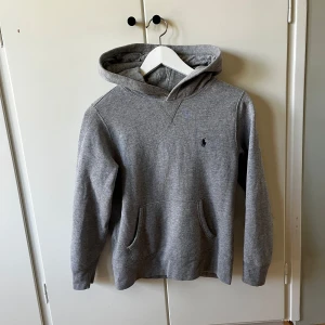 Grå hoodie från Polo Ralph Lauren - Klassisk grå hoodie från Polo Ralph Lauren med broderad logga på bröstet. Tröjan har huva, känguruficka och långa ärmar. Tillverkad i mjuk bomullsmix som är perfekt för chill dagar. Enkel och stilren design som funkar till allt.