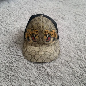 Gucci keps med tigertryck och GG-mönster - Snygg keps från Gucci med klassiskt GG-monogram i beige och brunt, prydd med två stora tigrar framtill. Baksidan har svart mesh och justerbart band med grönt och rött. Materialmix av bomull, polyester och polyuretan. Perfekt statement-piece för dig som gillar lyxiga accessoarer. 