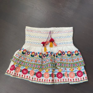 Vit broderad kortkjol med volang - Superfin vit kortkjol med färgglada broderade blommor och mönster. Kjolen har volang, smock i midjan och snörning med tofsar i rött, rosa och gult. Perfekt för sommarens alla äventyr och ger en bohemisk vibe. Från Zara 
