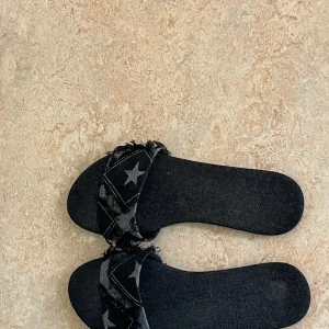 Svarta sandaler i denim med stjärnor - Coola svarta sandaler i denim med fransade kanter och stjärnmönster på remmen. Platt sula och öppen tå ger en avslappnad vibe. Perfekta för sommardagar när du vill sticka ut lite extra.