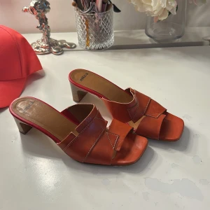 Röda sandaler med klack från Mitica - Snygga röda sandaler från Mitica med breda korsade remmar i skinn och kontrastsömmar. Modellen har fyrkantig öppen tå och låg klack i trälook. Perfekta för att ge en färgklick till din outfit. Insidan är fodrad i beige och röd ton.