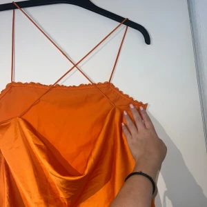 Orange satin topp med spets - Superfin orange topp i satin med tunna axelband som korsas i ryggen. Toppen har spetsdetaljer längs urringningen och en snygg drapering framtill. Perfekt för dig som vill sticka ut med färg och coola detaljer.  Passar s-l 