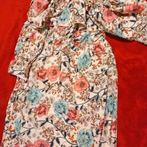 Blommig offshoulder klänning - Superfin kortklänning med blommigt mönster i pastellfärger. Offshoulder-modell med volang upptill och resår i midjan. Färgerna går i vitt, ljusblått, rosa och orange. Perfekt för sommarens alla tillfällen!