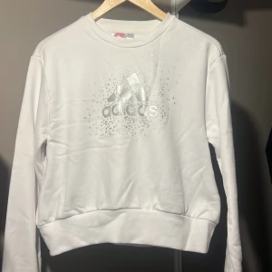 Vit sweatshirt med silvertryck från Adidas - Superfräsch vit sweatshirt från Adidas med glittrigt silvertryck och logga framtill. Tröjan har rund hals, långa ärmar och breda muddar. Perfekt för dig som gillar sportig och clean stil med lite extra bling.