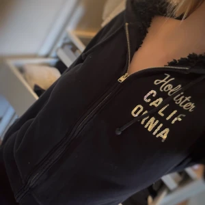 Marinblå zip hoodie från Hollister - Marinblå zip hoodie från Hollister med dragkedja framtill och broderad text i vitt och glittriga paljetter. Hoodien är fodrad!                                            Är i storlek M men passar S också!🩷                              Har liknande zip hoodies, kolla gärna in de! 