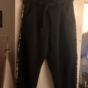 Svarta mjukisbyxor med leoparddetaljer - Svarta mjukisbyxor med leopardmönstrade revärer längs sidorna. Byxorna har resår i midjan och knytband framtill för extra komfort. Perfekta för en chill och trendig look. Mjukt material och praktiska fickor på sidorna.