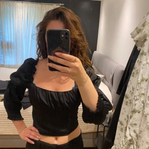 Svart puffärmad blus  - Supersnygg svart blus med puffiga trekvartsärmar och smockdetaljer vid ärmslut. Toppen har fyrkantig ringning och är gjord i ett glansigt, satininspirerat material. 