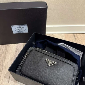 Svart axelväska från Prada i skinn (Byte✅) - Snygg svart axelväska från Prada i strukturerat skinn med silverfärgade dragkedjor och justerbar axelrem med logotyp. Väskan har två stora fack, ett mindre ytterfack och klassisk trekantig Prada-logga framtill. Perfekt för att bära det viktigaste med stil. Kvitto+Certifikat✅                                        Köper du den ny kostar den 29 000kr men jag säljer den för endast 15000kr runt 50% skillnad 