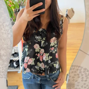 Svart blommig linnetopp med rosett - Supersöt svart linnetopp med blommigt mönster i rosa, vitt och grönt. Toppen har en liten rosett vid halsringningen och är ärmlös med smala axelband. Den är lätt och flowy, perfekt för varma dagar och ger en romantisk vibe.
