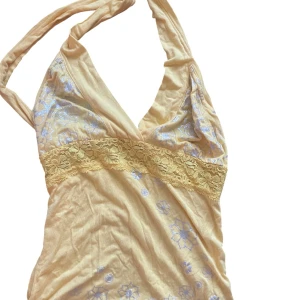 Gul blommig halterneck linnetopp - Söt gul halterneck linnetopp med silvriga blommor och spetsdetalj under bysten. Toppen har djup v-ringning och är ärmlös, perfekt för varma dagar. Materialet känns mjukt och lätt, och spetsen ger en extra touch. Passar dig som gillar färg och unika detaljer.