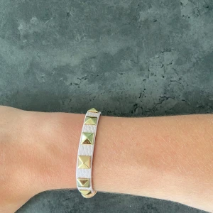 Valentino liknande armband  - Snyggt armband i ljusrosa skinnimitation med guldiga pyramidformade nitar längs hela bandet. Justerbar längd med flera hål och spänne i guldton. Perfekt accessoar för att ge outfiten lite edge.