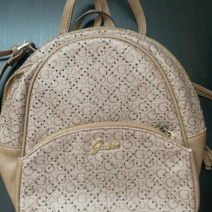 Beige ryggsäck från Guess med logomönster - Snygg beige ryggsäck från Guess med klassiskt logomönster över hela väskan och guldfärgade detaljer. Väskan har dragkedja, justerbara axelremmar och en praktisk ytterficka. Perfekt storlek för vardagens alla prylar och ger en trendig touch till din outfit.