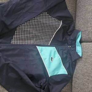 Lacoste tracksuit - Snygg vindjacka från Lacoste Sport i mörkblått med turkosa detaljer och vitt rutmönster på bröstet. Jackan har dragkedja framtill, hög krage och klassisk Lacoste-logga. Perfekt för dig som gillar sportig och stilren look.