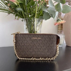 Brun kuvertväska Michael Kors - Snygg brun kuvertväska från Michael Kors med diskret mönster och guldfärgad kedja. Väskan har dragkedja upptill och logga i guld framtill. Perfekt storlek för det viktigaste och enkel att bära i handen eller på axeln. Den är helt ny.