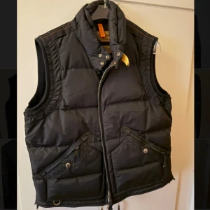 Svart dunväst från Parajumpers - Snygg svart dunväst från Parajumpers med hög krage, dragkedja och tryckknappar framtill. Västen har två stora fickor med dragkedja och knapp, samt coola detaljer som orange rem och logga i nacken. Perfekt för lager-på-lager-stil.