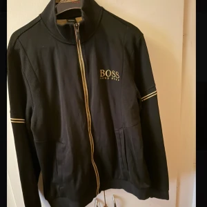 Svart zip tröja från Hugo Boss - Svart zip tröja från Hugo Boss med gulddetaljer och dragkedja framtill. Tröjan har ribbade partier på axlar och ärmar samt diskreta ränder på ärmarna. Klassisk ståkrage och BOSS-logga på bröstet ger en clean och sportig vibe.
