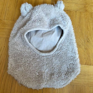 Beige fluffig balaclava med öron H&M - Supermjuk och fluffig balaclava från H&M i beige färg, med söta små öron upptill. Utsidan är pälsliknande och insidan är fodrad med bomull för extra komfort. Perfekt för kalla dagar och ger en gullig look.