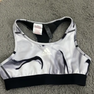 Adidas sporttopp i svart och vitt - Sportig topp från Adidas i svart och vitt med abstrakt mönster och logga framtill. Modellen är ärmlös med brottarrygg och bred resår nedtill. Tillverkad i ett stretchigt syntetmaterial som andas, perfekt för träning och aktivitet.