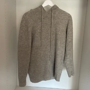 Grå stickad hoodie med huva - Mysig grå stickad hoodie med huva och snörning. Tröjan har lång ärm och ribbade muddar vid ärmslut och nederkant. Perfekt för lager-på-lager och chill dagar. Enkel och stilren design som funkar till det mesta.