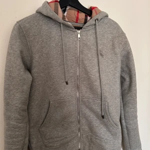 Grå hoodie från Burberry med dragkedja - Snygg grå hoodie från Burberry med klassiskt rutigt foder i huvan. Tröjan har dragkedja framtill, snörning i huvan och två stora fickor. Perfekt för dig som gillar stilrena plagg med en touch av lyx. Äkta såklart! Hör av dig vid funderingar! 