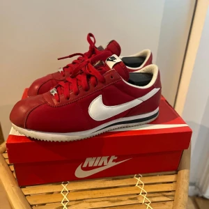 Nike Cortez röd/vit sneakers - Snygga Nike Cortez sneakers i rött med vita detaljer och klassisk vit swoosh på sidorna. Skorna har snörning, låg profil och platt sula. Ovandelen är i skinn och syntet, med vit och svart rand på mellansulan. Perfekta för dig som gillar retrostil.