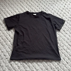 Svart t-shirt med pärldetaljer Zara - Svart t-shirt från Zara i bomull med korta ärmar. Ärmsluten är dekorerade med snygga pärldetaljer som ger en cool touch. Klassisk rak passform och rund halsringning!💕