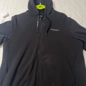 Svart hoodie från Armani Exchange - Svart hoodie från Armani Exchange med dragkedja framtill och justerbar huva med snörning. Tröjan har diskret logga på bröstet och är tillverkad i mjuk bomullsblandning. Perfekt för en chill och stilren look.