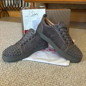 Grå mocka sneakers från Louboutin - Snygga grå sneakers i mocka från Christian Louboutin med ikoniska röda sulor och coola nitar på tån. Skorna har snörning och en rund tå, samt en platt sula. Perfekta för dig som vill sticka ut med exklusiva detaljer och edgy vibe.