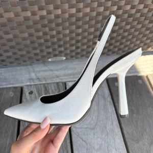 Vita pumps med klack från Bershka - Snygga vita pumps från Bershka i storlek 39. Skorna har spetsig tå, öppen häl med rem och hög klack. Aldrig använda