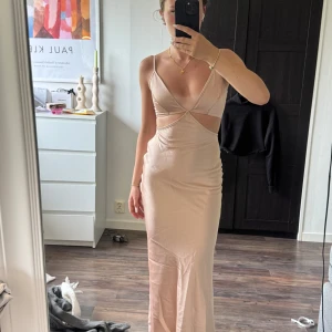 Champagnefärgad långklänning med cut-outs - Supersnygg långklänning i champagnefärg med smala axelband, djup v-ringning och trendiga cut-outs vid midjan. Klänningen har figurnära passform och släpper ut lätt nertill. Perfekt för dig som vill sticka ut med en elegant och modern look.