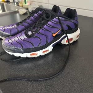 Nike Air Max Plus Tn lila och svart - Nike Air Max Plus Tn sneakers i lila och svart med orange swoosh och detaljer. Skorna har en vågig design på ovandelen, vit sula med synliga luftbubblor och svarta snören. Snygg streetstil med unika färgkombinationer och Tn-logo på hälen.