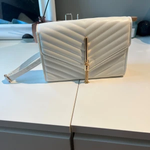 Vit quiltad axelväska med gulddetalj - Snygg vit axelväska med quiltat mönster och guldfärgad kedjedetalj framtill. Väskan har en stilren, rektangulär form och justerbart axelband. Perfekt för att lyfta din outfit med en elegant touch.