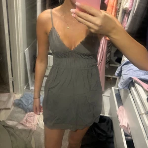 Grå kort klänning med smala axelband - Supersöt grå kort klänning med smala axelband och v-ringning. Klänningen har en markerad midja och lätt rynkad kjol, perfekt för varma dagar. Enkel och stilren design som är lätt att matcha med accessoarer.