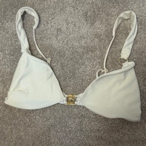 Vit bikiniöverdel med gulddetalj - Snygg vit bikiniöverdel med trekantsformade kupor och justerbara axelband. Framtill finns ett elegant guldfärgat spänne som ger en exklusiv touch. Materialet är glansigt och stretchigt, perfekt för strandhäng eller poolparty.