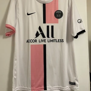 PSG Nike vit och rosa fotbollströja - Paris Saint-Germain fotbollströja från Nike i vitt med rosa och svart vertikal rand på framsidan. PSG-logga på bröstet, svart Nike-swoosh och sponsortryck. Jätte fint material och den är i perfekt skick! 