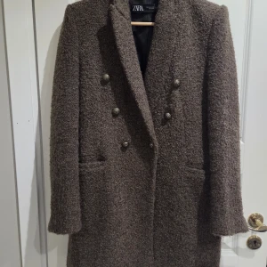 Brun kappa från Zara med dubbelknäppning - Säljer en brun kappa från Zara med snygg dubbelknäppning och grov textur. Jackan har klassisk krage, två fickor framtill och långa ärmar. Perfekt för dig som vill ha en stilren och trendig look under kallare dagar.