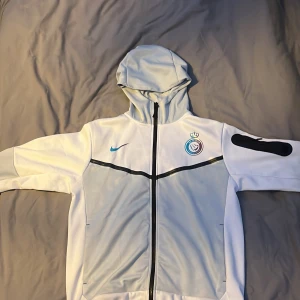 Nike vit och ljusgrå - Nike i vitt och ljusblått med huva och hel dragkedja. Jackan har svart Nike-logga på ärmen, klubbmärke på bröstet och blå Nike swoosh. Tillverkad i 100% polyester, perfekt för dig som gillar sportig stil. Passar också perfekt om du är Ronaldo fan👍