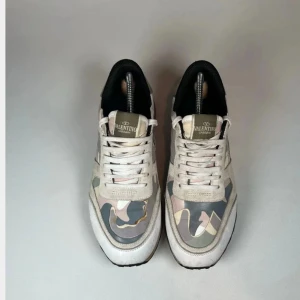 Valentino Rockrunner sneakers camo - Snygga Valentino Rockrunner sneakers med unik camouflagemönstrad ovandel i beige, rosa, grått och vitt. Skorna har detaljer i mocka, skinn och textil, samt guldigt hälparti och nitar bak. Klassisk snörning och svart yttersula för en cool och lyxig vibe.